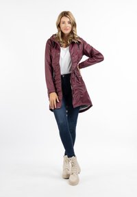 Jachetă waterproof de culoare vinie cu glugă, talie strânsă și închidere cu nasturi. Puritată peste un top alb și blugi albaștri, asortată cu botine bej.