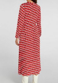 Robe rouge à manches longues avec un motif géométrique de formes blanches et rouge plus foncé, présentant une taille cintrée et une jupe fluide.