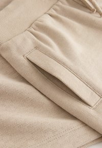 Gros plan sur un pantalon de survêtement beige montrant une taille élastique et une poche latérale fendue avec des coutures détaillées sur un tissu texturé.