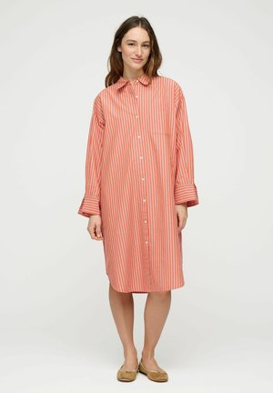 Ženska, ki nosi koralno in belo dolgo srajčno obleko z navpičnimi črtami ter bež balerinke, stoji pred enobarvnim ozadjem.