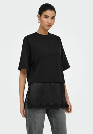 Kvinne iført en svart oversized kortermet topp lagd over en svart blonderkantet camisole, kombinert med grå jeans, vendt mot front.