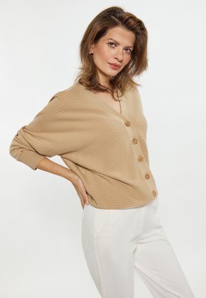 Beige geribbelde cardigan met een V-hals, voorzien van zes houten knopen aan de voorkant. Draagt over witte broek, met ontspannen mouwen.