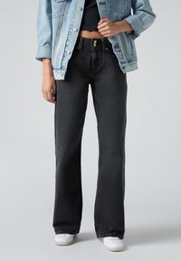 Personne portant un jean large foncé, des baskets blanches, un top court noir et une veste en jean bleu clair sur un fond uni.