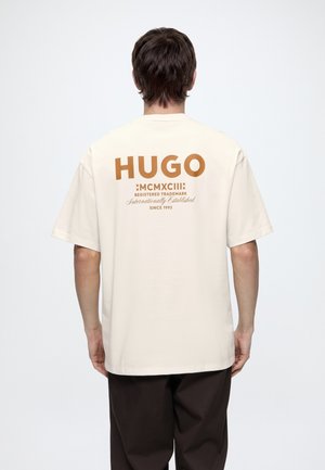 Homme vu de dos portant un T-shirt crème avec un grand texte marron "HUGO MCMXCIII" et un pantalon noir sur un fond blanc uni.