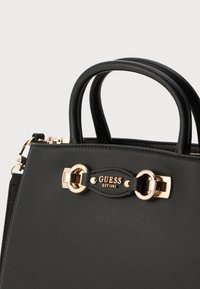 Sac en cuir noir avec des accents dorés. Présente deux poignées et une plaque décorative affichant "GUESS EST 1981". Texture lisse.