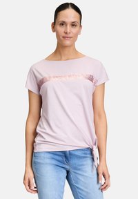 Jasnoróżowa koszulka z krótkim rękawem, z cekinowym poziomym paskiem na wysokości klatki piersiowej oraz detalem w postaci zawiązania z boku. Miękka tkanina, luźny krój.