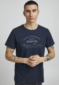 Tamno plavi pamučni majica s okruglim izrezom, s teksturiranim materijalom i otisnutim tekstom u vintage stilu preko prsa.