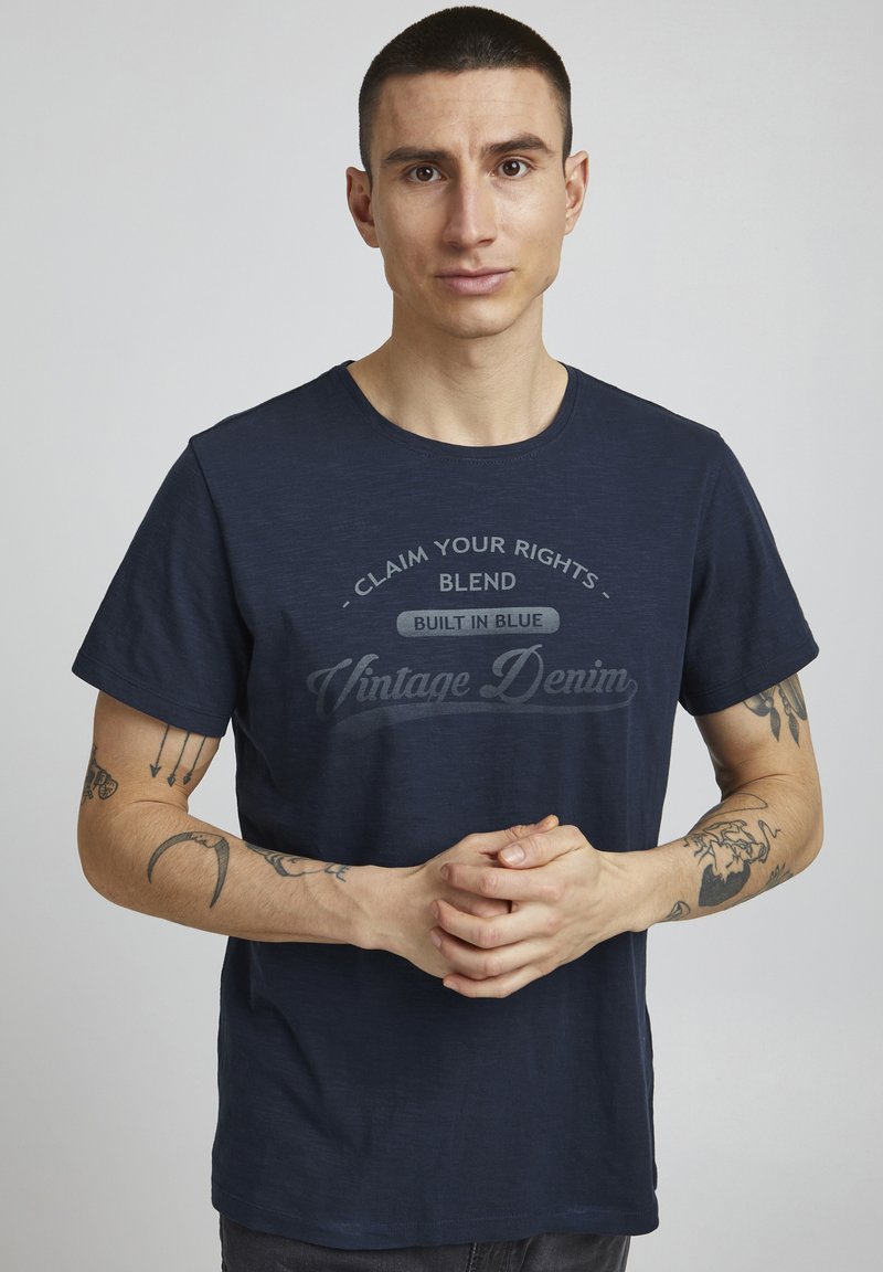Tamno plavi pamučni majica s okruglim izrezom, s teksturiranim materijalom i otisnutim tekstom u vintage stilu preko prsa.