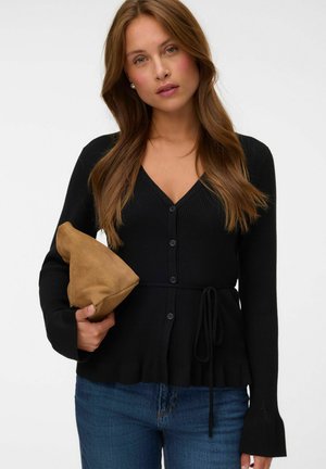 Woman with long brown hair wearing a black buttoned cardigan and blue jeans, holding a folded tan suede clutch bag against a white background.