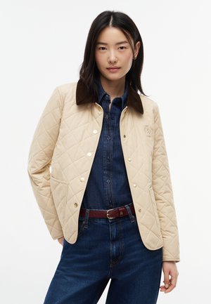Kvinde med glat sort hår iført beige quiltet jakke over mørkeblå denimskjorte og jeans med brunt bælte, står mod hvid baggrund.