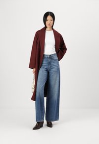 Abrigo largo burdeos, blusa blanca ajustada, vaqueros de pierna ancha azules, botas de tobillo marrones con tacón y un bolso pequeño claro. Tejidos suaves, diseño a medida.