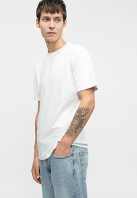 Biała bawełniana koszulka z okrągłym dekoltem, krótkimi rękawami i luźnym krojem, w połączeniu z jasnymi dżinsami. Zauważalny tatuaż na przedramieniu.