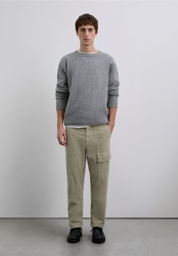 Suéter gris de punto, ajuste holgado, puños acanalados; pantalones cargo verde claro con bolsillos laterales; zapatos negros con cordones; fondo liso.