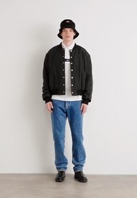 Giacca bomber nera, polo bianca, jeans blu e stivali neri. Uomo che indossa un cappello a secchiello nero con un logo. Outfit semplice e casual.