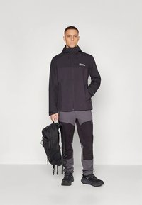 Giacca impermeabile nera con cerniera frontale, dettagli grigi e cappuccio; abbinata a pantaloni grigi e neri. Modello con zaino nero in mano.