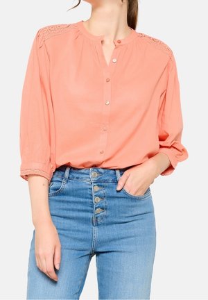 Vrouw draagt een zalmroze blouse met knopen en kantdetails op de schouders, gecombineerd met blauwe jeans met hoge taille die vier zichtbare knopen hebben.