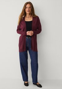 Bordeaux cardigan met open voorkant, lange mouwen en een gladde textuur, gecombineerd met een zwarte top en blauwe wijde jeans. Zwarte ballerina's.