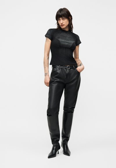 Γυναίκα με σκούρα μαλλιά που φοράει εφαρμοστή μαύρη υφασμάτινη μπλούζα με κοντό μανίκι, μαύρο δερμάτινο παντελόνι με ζώνη και μαύρες μπότες μέχρι το γόνατο, στέκεται με το χέρι στην τσέπη.