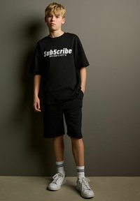 Sort oversized T-shirt med hvid tekst, parret med sorte shorts. Hvide sneakers med gråstribede sokker fuldender det afslappede outfit.