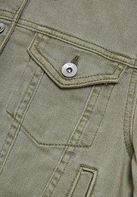 Grön denimjacka fragment med en knäppt ficklock, synlig sömnad och en metallknapp med "EST 1844" ingraverad.