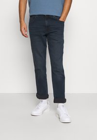 Jeans de mezclilla de un azul oscuro con puños doblados, combinados con zapatillas blancas. El conjunto presenta un diseño sencillo con un corte ajustado.