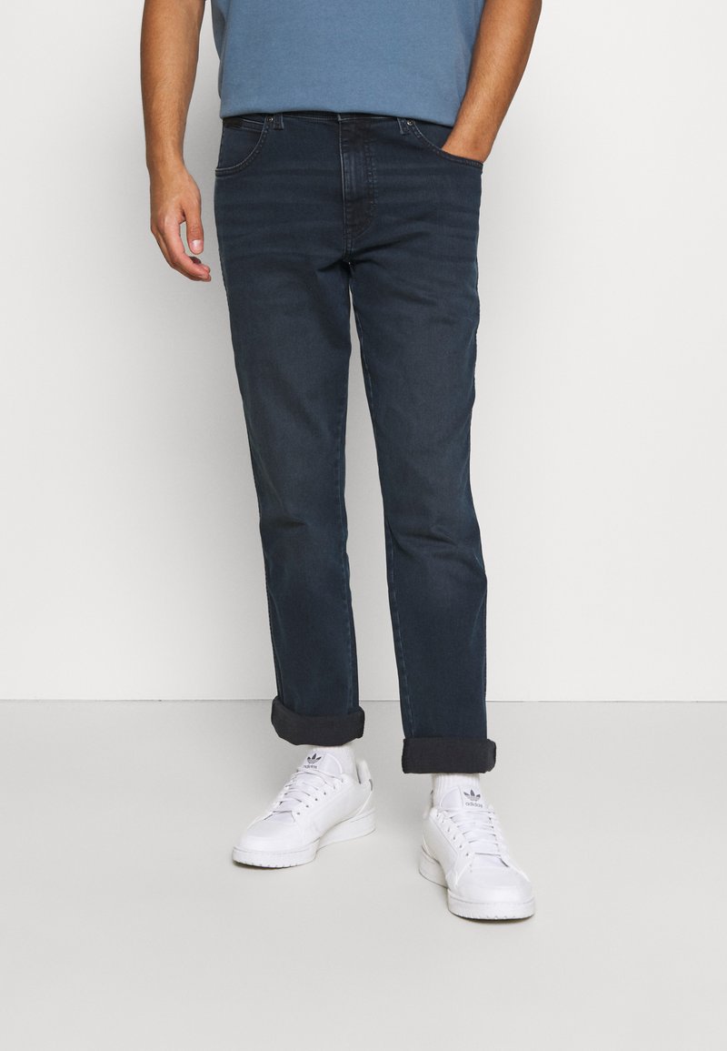 Jeans de mezclilla de un azul oscuro con puños doblados, combinados con zapatillas blancas. El conjunto presenta un diseño sencillo con un corte ajustado.