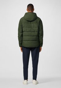 Chaqueta acolchada verde con costuras horizontales, capucha adjunta y un logo en la parte de atrás, llevada sobre joggers azul marino y zapatillas blancas.