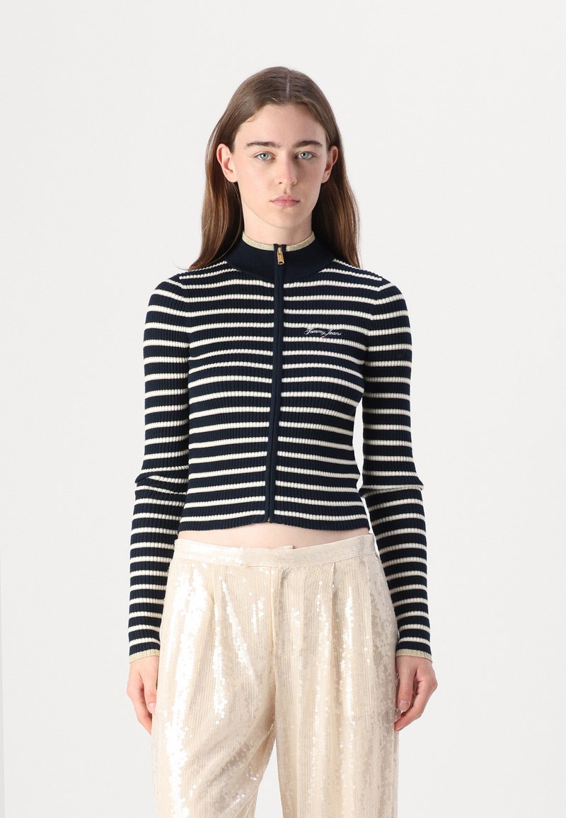 Navy and cream striped ribbed zip-up cardigan with a high collar and gold zipper, paired with light sequined wide-leg pants.