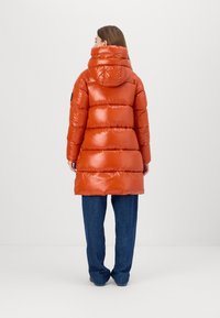 Puffer coat in shiny orange with a high neck and hood, featuring a segmented design. Worn with loose-fitting blue jeans.