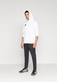 Λευκή φούτερ με κουκούλα και τσέπη καγκουρό, με μαύρο λογότυπο, σε συνδυασμό με σκούρο γκρι παντελόνια φόρμας με υφή και μαύρα αθλητικά παπούτσια.