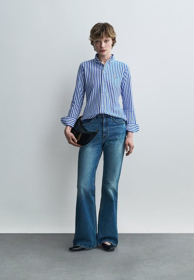 Γυναίκα που φοράει μπλε και λευκό πουκάμισο με κάθετες ρίγες και κουμπιά, μπλε τζιν καμπάνα, μαύρα παπούτσια, κρατώντας μαύρο τσαντάκι, στέκεται μπροστά σε ανοιχτό γκρι τοίχο.