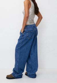 Losse blauwe denim jeans met een hoge taille en twee achterzakken, gecombineerd met een licht sprankelend grijs tanktop en bruine klompen.