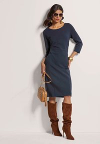 Marinefarbendes, figurbetontes Kleid mit Dreiviertelärmeln, kombiniert mit braunen Wildleder-Stiefeln bis zum Knie und einer hellbraun gequilteten Handtasche. Die Accessoires umfassen Ohrhänger.