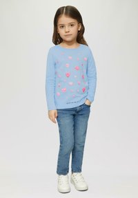 Chemise à manches longues bleu clair avec des motifs de cœurs roses ; associée à un jean bleu et des baskets blanches. Le texte en bas dit "Répandre l'amour et la joie".