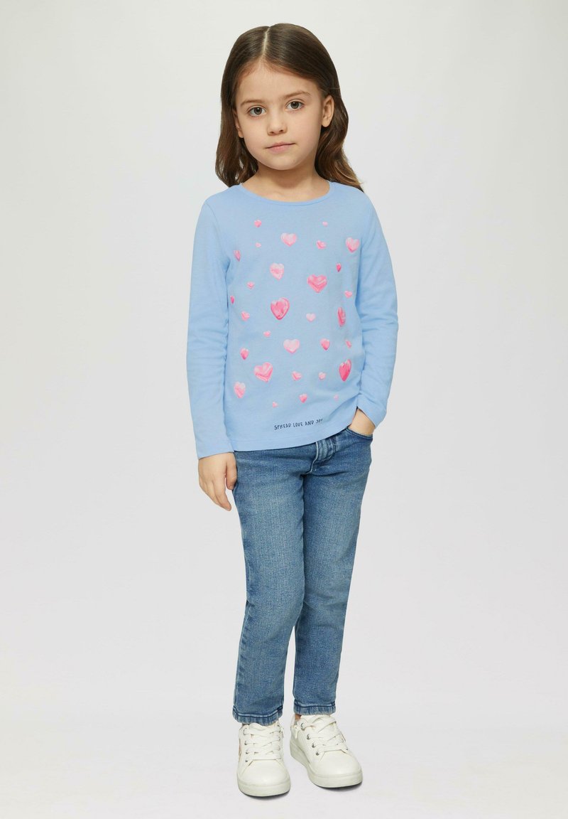 Chemise à manches longues bleu clair avec des motifs de cœurs roses ; associée à un jean bleu et des baskets blanches. Le texte en bas dit "Répandre l'amour et la joie".