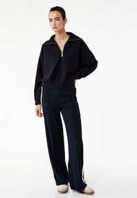 Felpa zippata blu marina con colletto alto e maniche ampie, abbinata a pantaloni larghi scuri con strisce laterali crema.