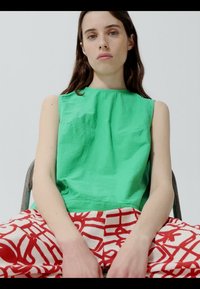 Jeune femme aux longs cheveux bruns assise, portant un haut vert sans manches et un pantalon à motifs rouges et blancs, sur un fond uni.