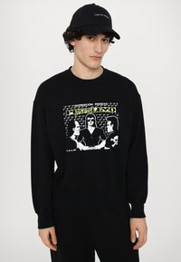 Black sweatshirt with a graphic print featuring three figures, surrounded by a text banner, and ribbed cuffs and hem. Worn with a matching black cap.