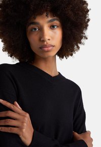 Młoda osoba o kręconych włosach w czarnym swetrze z dekoltem w serek, z założonymi rękami, patrząca prosto przed siebie na jednolitym tle.