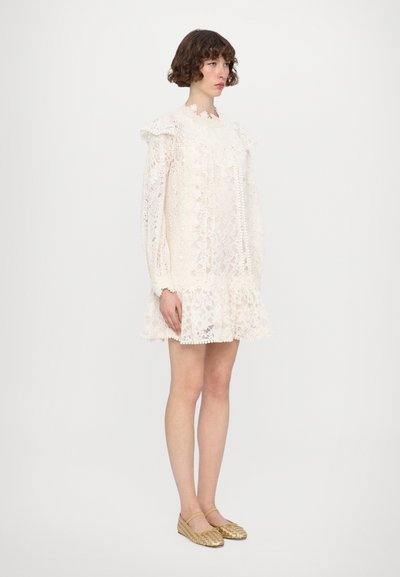 Kleid aus Spitze in Cremefarbe mit langen Ärmeln und Rüschenausschnitt. Verfügt über einen ausgestellten Saum. Kombiniert mit gewebten goldenen Ballerinas.