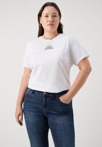 Mujer vistiendo una camiseta blanca con un gráfico de sol y el texto "La vida es buena", combinada con jeans oscuros, de pie con una mano en el bolsillo.