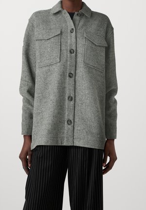 Veste en laine grise boutonnée avec deux poches avant, portée sur un pantalon noir à fines rayures, sur une personne debout.