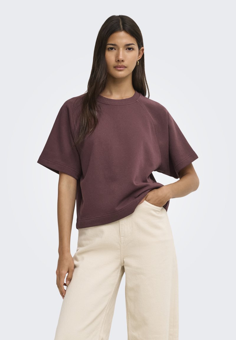 Jeune femme aux longs cheveux foncés portant un t-shirt ample violet foncé à manches courtes et un pantalon beige clair, debout avec une main dans la poche.