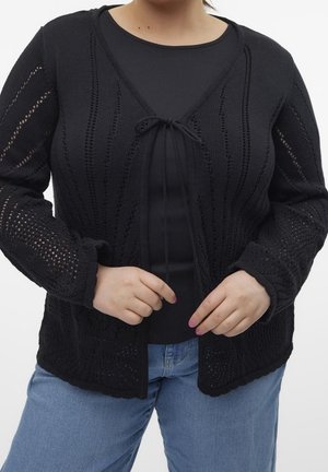 Mustan neuleliivin avoimella designilla, jossa on solmimisella suljenta, teksturoituja kuvioita ja pitkät hihat, kerroksittain mustan paidan päällä.