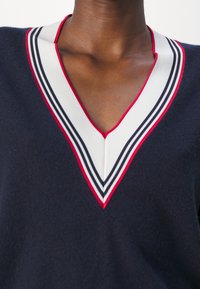 Pull bleu marine foncé avec col en V blanc bordé de rayures rouges et noires, porté par une personne avec un cou et le haut de la poitrine visibles.