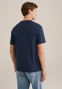 Moški obrnjena stran v enostavni temno modri majici s kratkimi rokavi in svetlih modrih kavbojkah na nevtralni podlagi.