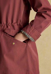 Bordowa kurtka z marszczonym paskiem w talii, z kieszenią zapinaną na mosiężny zatrzask i podszewką mankietów w kratę. Widoczna nazwa marki "Joules".