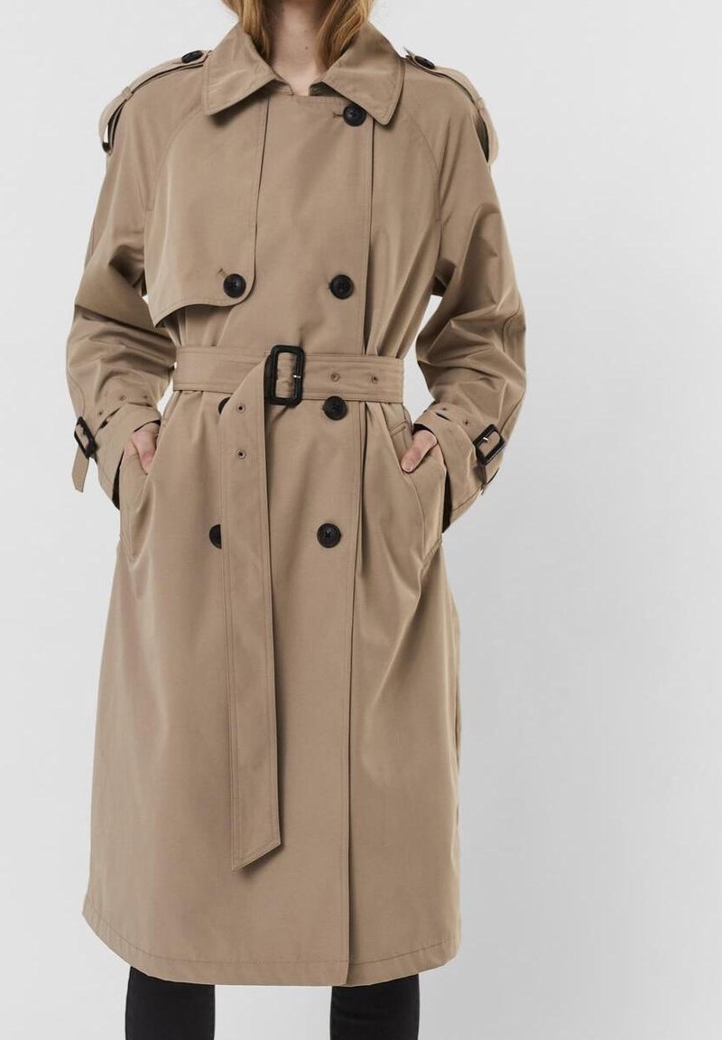 Femme portant un trench-coat beige croisé avec ceinture à la taille et boutons noirs, mains dans les poches, sur un fond clair uni.