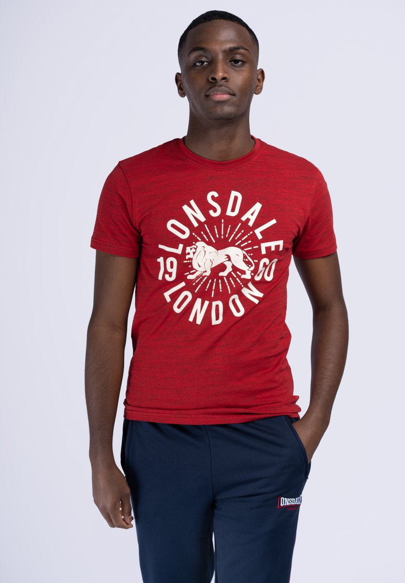 Rød t-shirt med korte ærmer, der har et hvidt grafisk print med en løve, ordene "Lonsdale London" og et vintage-år i cirkulært design.