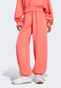 Korálové tepláky s uvoľneným strihom, elastickým pásom a manžetami na členkoch; s embosovaným logom Adidas. Skombinované s bielymi teniskami.